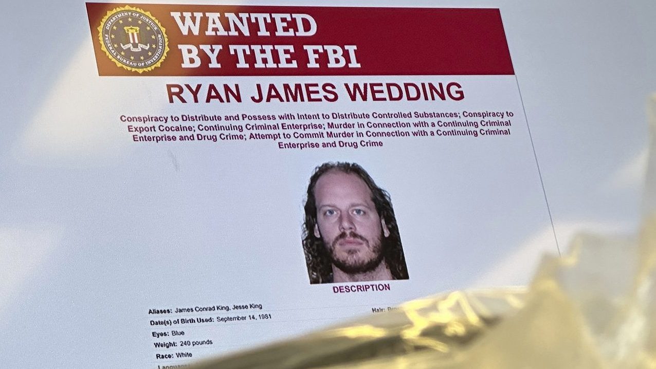 An image of former Canadian Olympic snowboarder Ryan Wedding, who is a fugitive and been charged with allegedly running and participating in a transnational drug trafficking operation, is displayed on a video monitor along with bricks of cocaine, foreground, during a news conference at the FBI offices in Los Angeles on Oct. 17, 2024. (AP Photo/Damian Dovarganes, File)